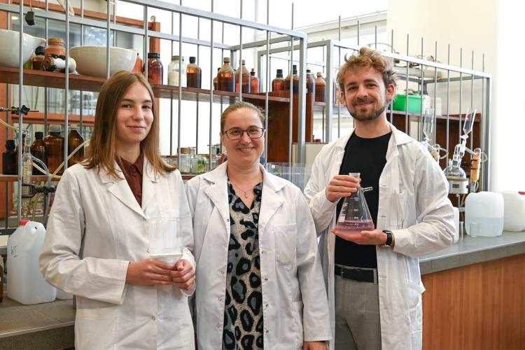 Na zdjęciu trzy osoby: dwie kobiety i mężczyzna, ubrani w fartuchy, stoją w laboratorium chemicznym