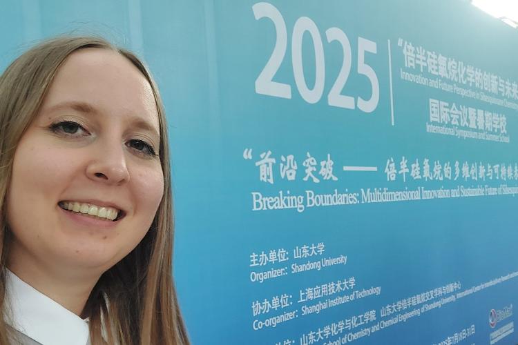 Na zdjęciu mgr inż. Anna Łabęda na tle niebieskiej ściany z infromacjami o konferencji i szkole letniej w Chinach