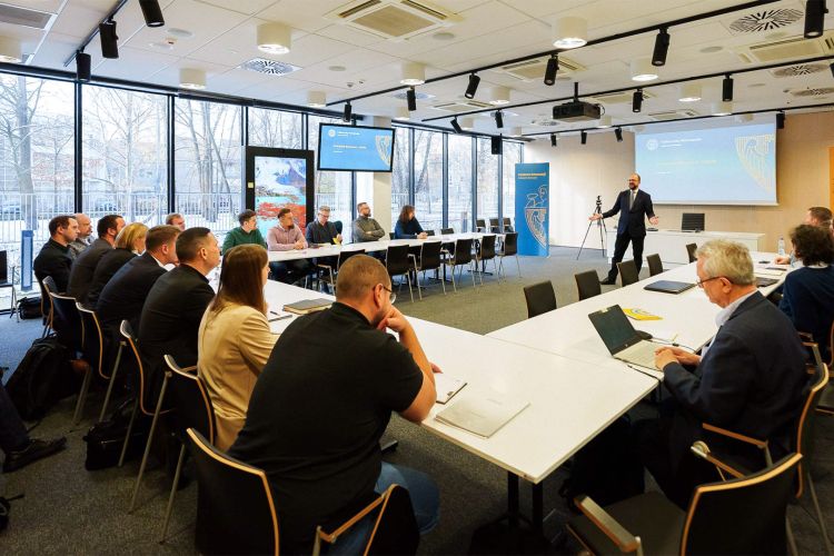 Na zdjęciu ludzie siedzący w sali przy stole w kształcie litery U. Z przodu tablica oraz prezentujący temat mężczyzna