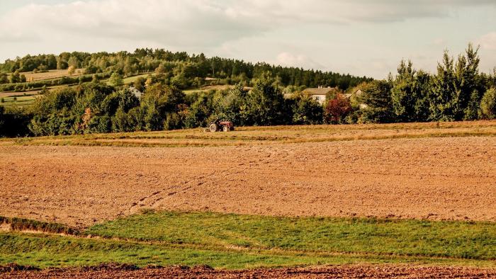 Na zdjęciu wiejski krajobraz: na pierwszym planie pole, w tle ciągnik, las i dom