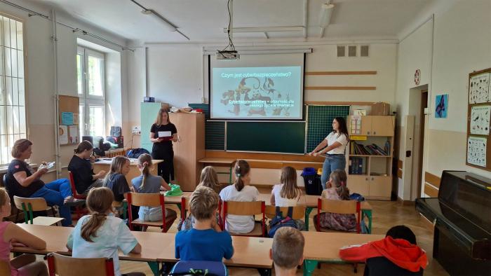 Na zdjęciu sala szkolna wypełniona dziećmi, które siedzą w ławkach. Z przodu dwie studentki stoją przy tablicy i prowadzą zajęcia