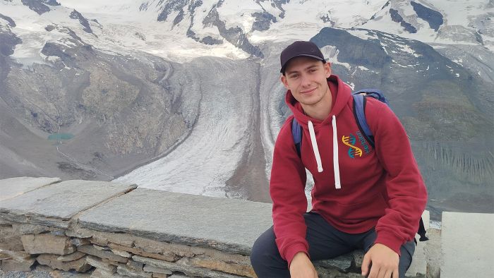 Na zdjęciu młody mężczyzna siedzi na murku, w tle widać góry - Alpy
