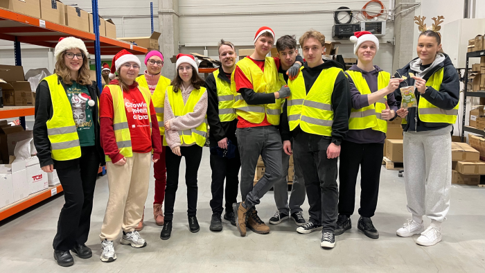 Zdjęcie przedstawia grupę 10 studentów, członków Studenckiego Koła Logistyki Stosowanej. Ubrani są w odblaskowe kamizelki, część z nich ma na sobie czapki świąteczne. Studenci stoją w magazynie między regałami z kartonami podczas wolontariatu realizowanego w ramach akcji Szlachetna Paczka.