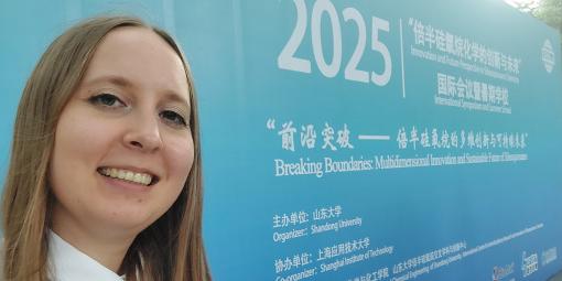 Na zdjęciu mgr inż. Anna Łabęda na tle niebieskiej ściany z infromacjami o konferencji i szkole letniej w Chinach