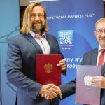 Na zdjęciu Marcin Stanecki – Główny Inspektor Pracy oraz prof. dr hab. inż. Andrzej Garbacz – Dziekan Wydziału Inżynierii Lądowej PW stoją na tle ścianek i ściskają sobie dłonie