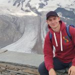 Na zdjęciu młody mężczyzna siedzi na murku, w tle widać góry - Alpy