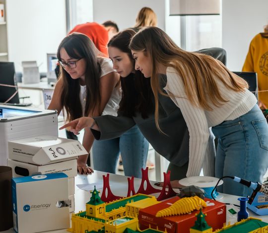 Na zdjęciu trzy studentki pochylają się nad drukarką 3D